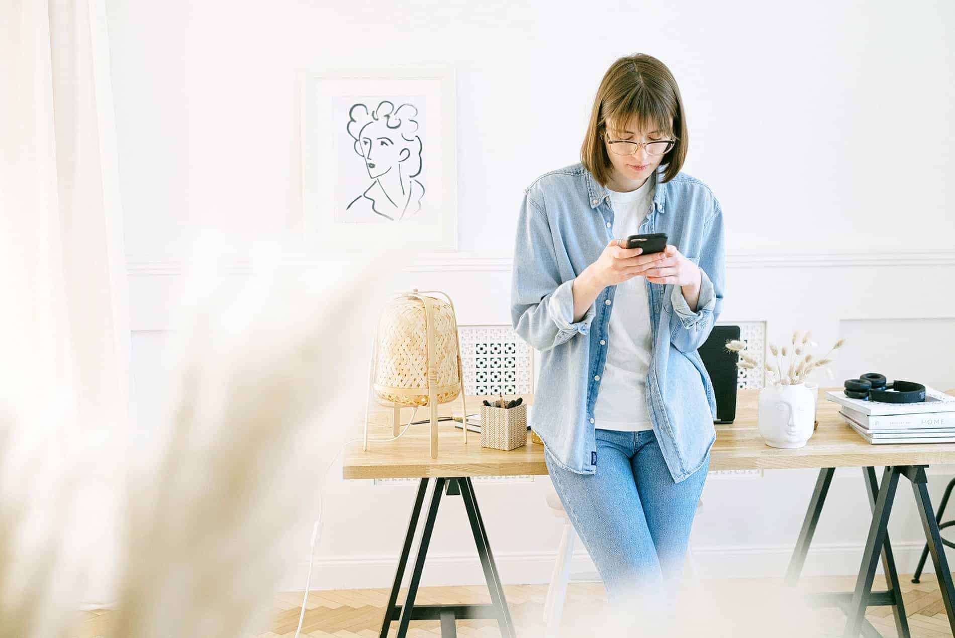 A person in a denim shirt uses a phone while leaning on a desk, with minimal decor in a bright, modern room.