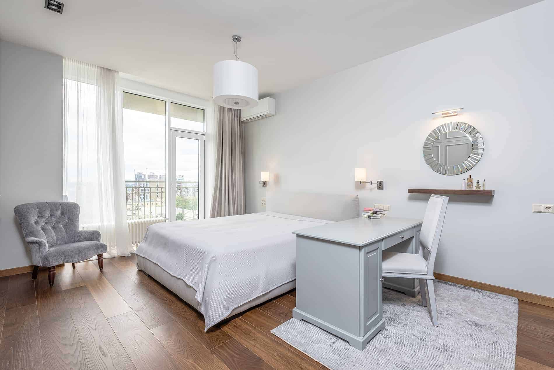 Bright bedroom with white walls, wooden floor, large bed, desk, chair, armchair, mirror, and balcony view of cityscape. Minimalist and modern design.