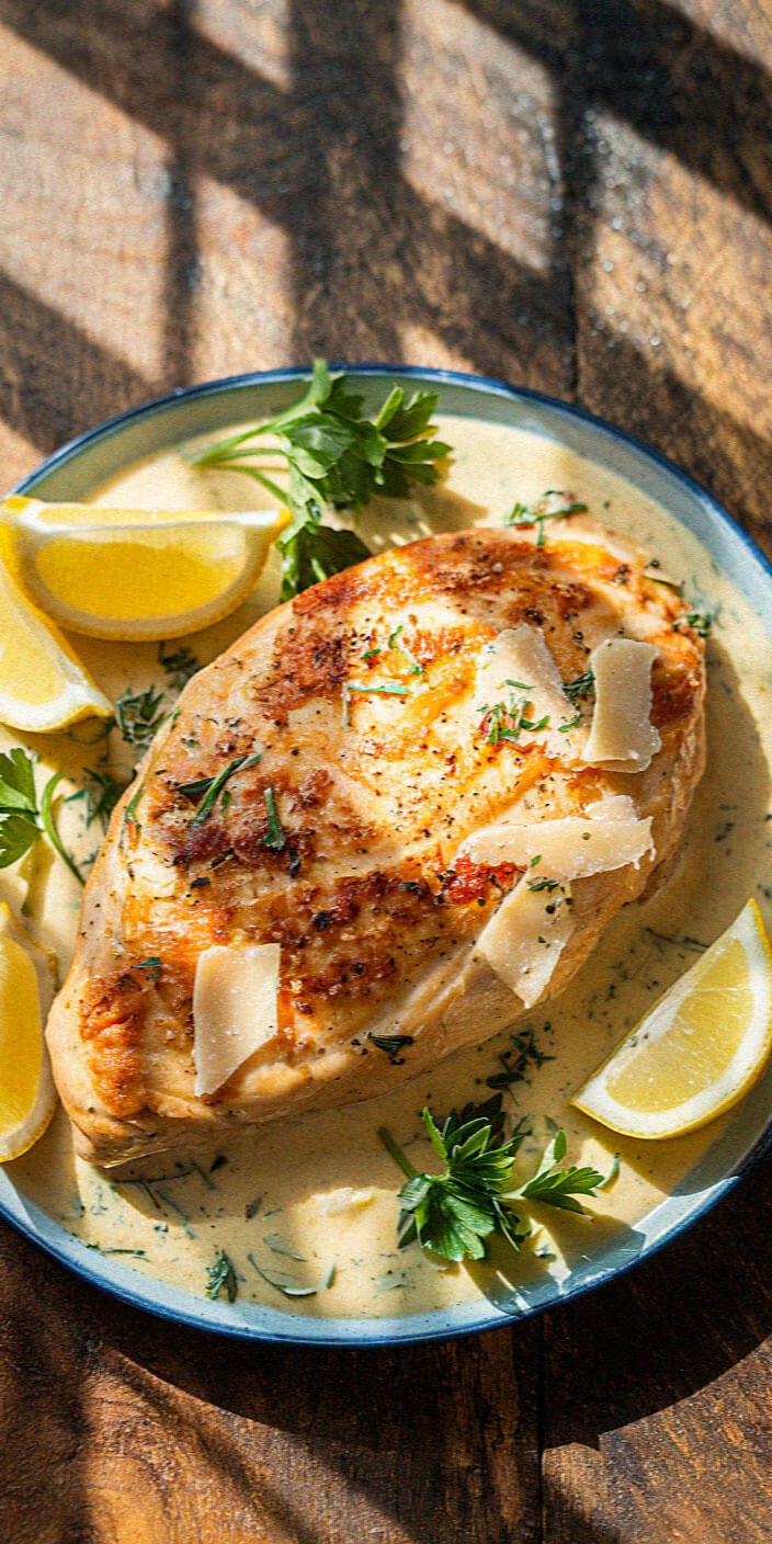 Grilled chicken breast on creamy sauce, garnished with lemon wedges, herbs, and parmesan shavings; served on a rustic wooden table.