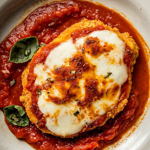 A plate of chicken parmesan with melted cheese and tomato sauce, garnished with fresh basil leaves, presented on a white dish.
