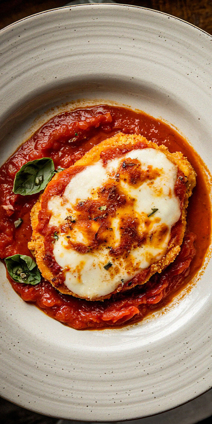 A plate of chicken parmesan with melted cheese and tomato sauce, garnished with fresh basil leaves, presented on a white dish.