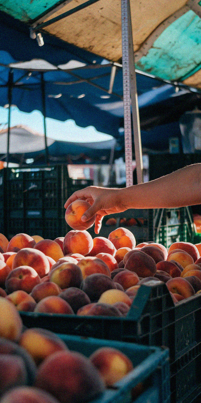 A person's hand selects a peach from a pile at an outdoor market stand, shaded by colorful tarps overhead.
