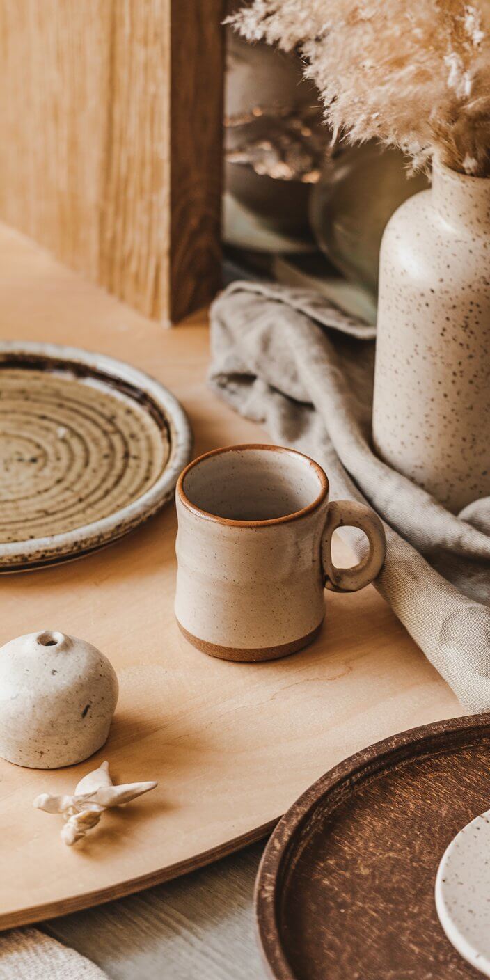 Ceramic cup, plate, and vase on a wooden surface with neutral colors and dried flowers creating a cozy, minimalist aesthetic.