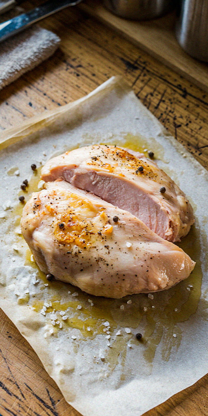 Two seasoned chicken breasts on parchment paper, sprinkled with salt and pepper, resting on a rustic wooden table, ready for cooking.