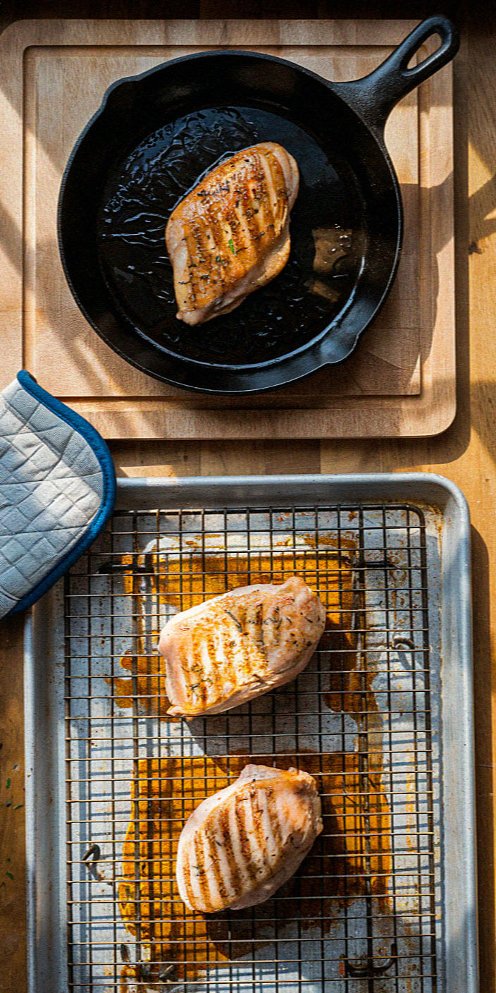 Three grilled chicken breasts: one in a cast iron skillet, two on a baking tray with a wire rack and oven mitt nearby.