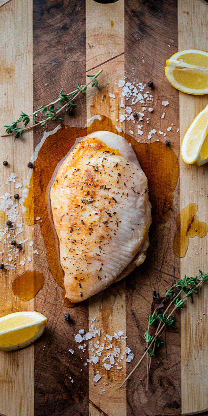 Grilled chicken breast seasoned with herbs, accompanied by lemon wedges, salt, peppercorns, and fresh thyme, displayed on a wooden cutting board.