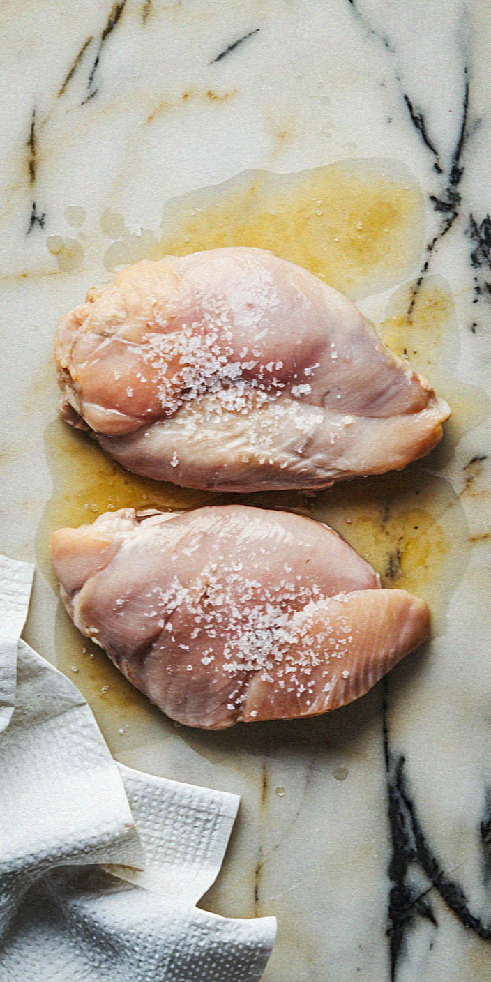 Two raw chicken breasts with salt on a marble surface. A paper towel is placed nearby.