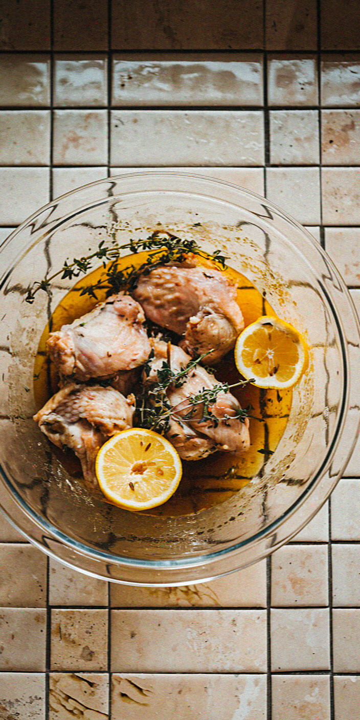 A bowl with marinated chicken, lemon slices, and herbs on a tiled kitchen countertop, ready for cooking.