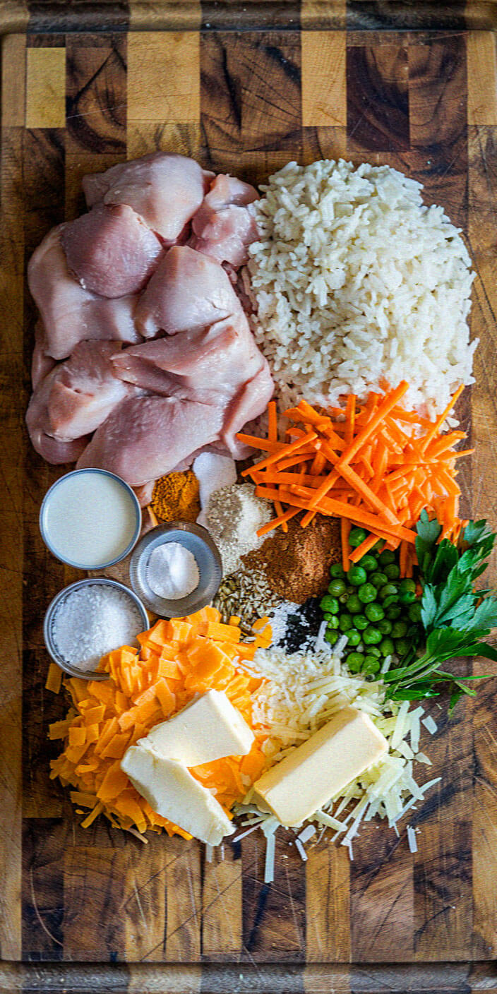 Raw chicken, rice, carrots, peas, cheese, butter, herbs, spices, and salt arranged on a wooden cutting board, ready for meal preparation.