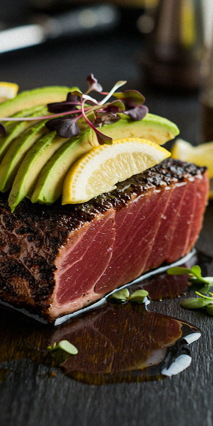 A beautifully plated dish featuring seared tuna topped with avocado slices, lemon wedge, and microgreens on a dark, stylish background.