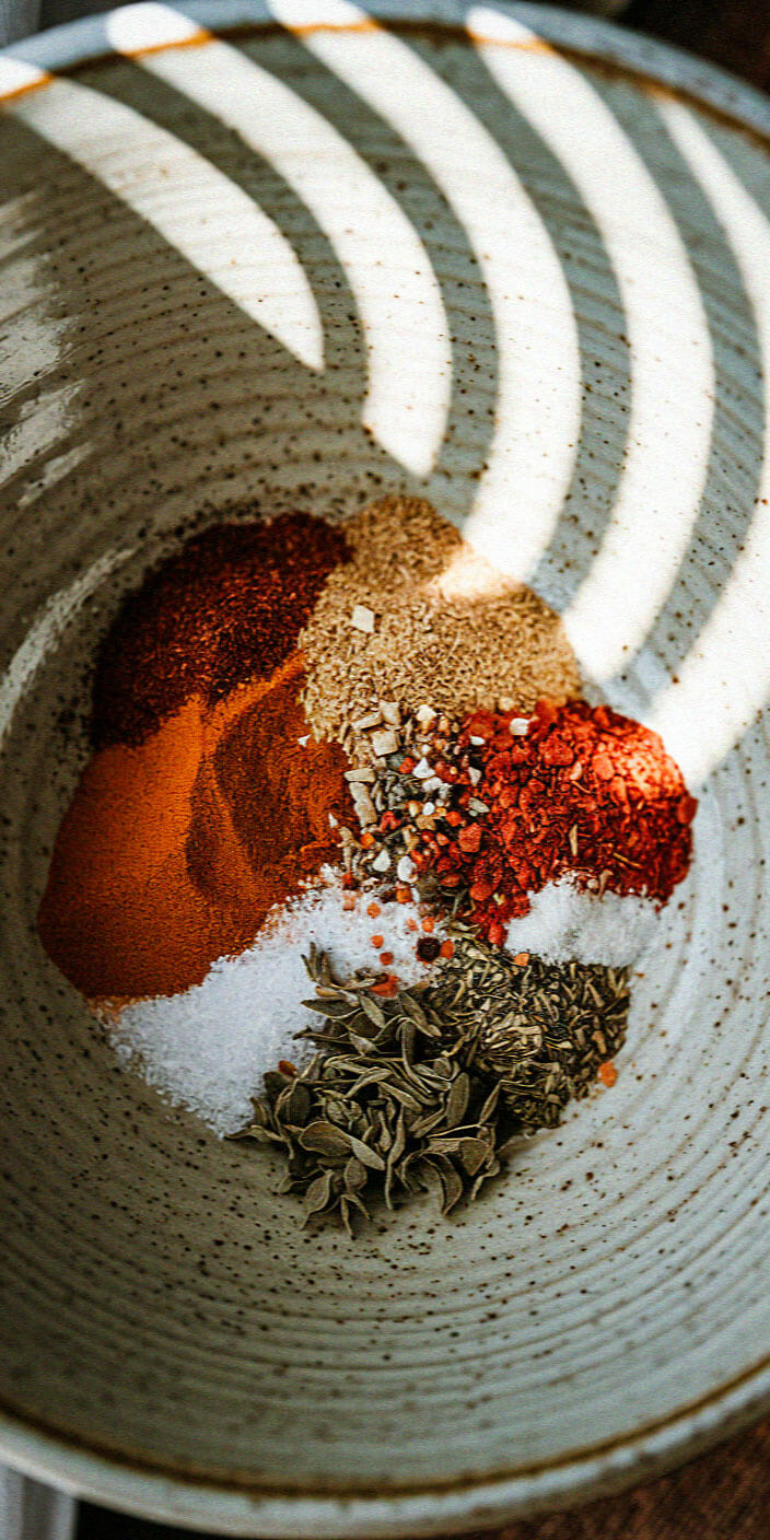 A ceramic bowl filled with colorful spices, including paprika, salt, and dried herbs, is illuminated by sunlight patterns.