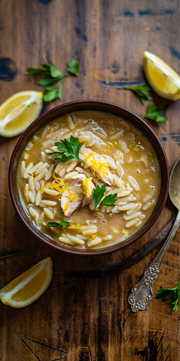 A bowl of orzo soup garnished with lemon zest and parsley, placed on a wooden table alongside lemon wedges and an ornate silver spoon.