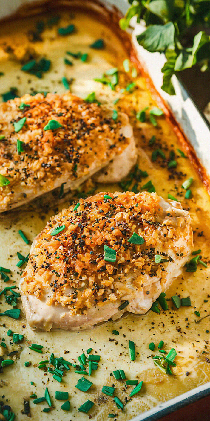 Close-up of seasoned baked chicken breasts, garnished with chopped herbs and crispy topping, in a baking dish with fresh greens nearby.