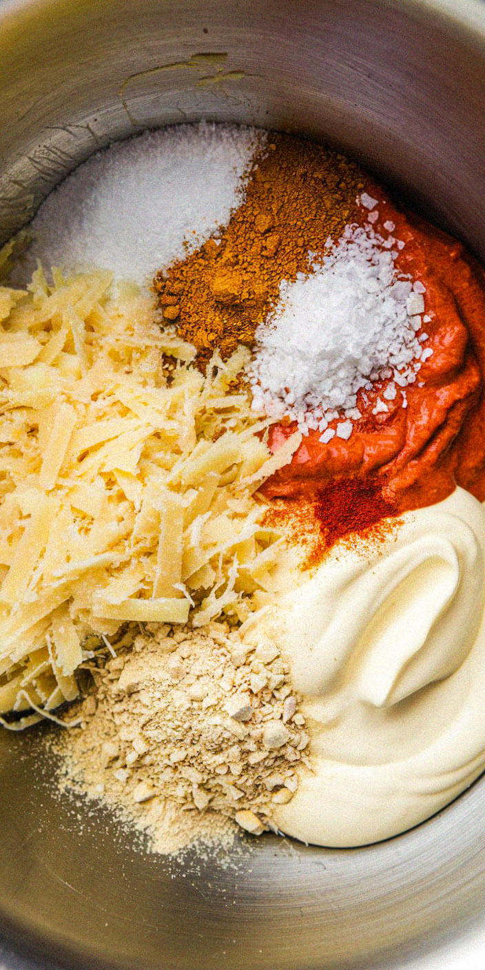 A mixing bowl contains grated cheese, spices, mayonnaise, and crushed nuts, ready for blending into a flavorful mixture.