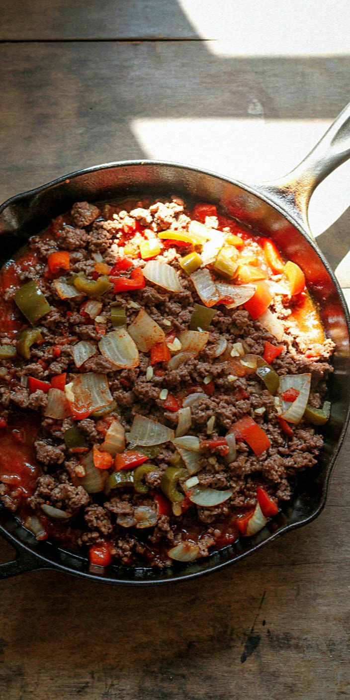 A cast iron skillet on a wooden table contains a hearty mix of ground meat, onions, and bell peppers in tomato sauce.