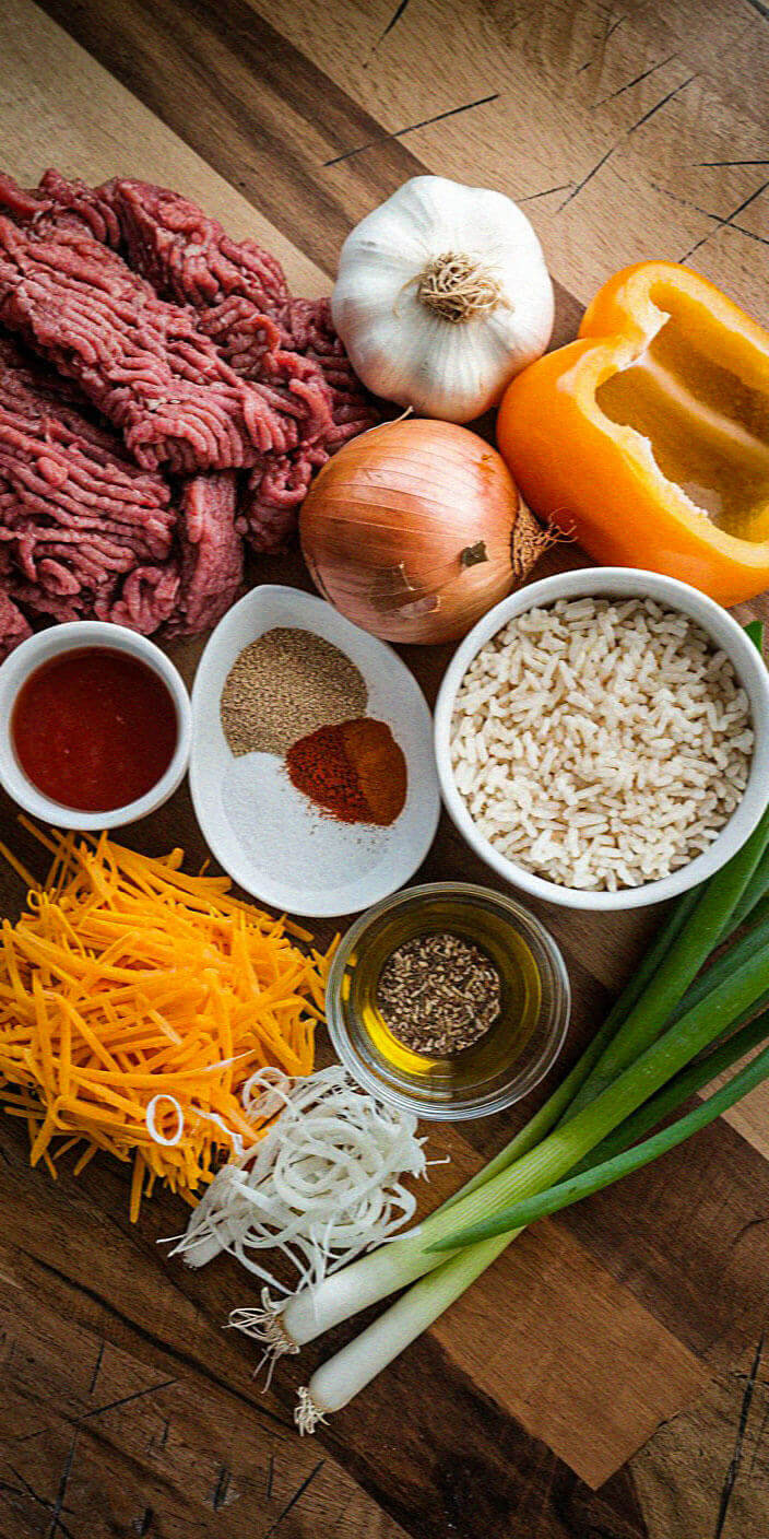 Ingredients for a dish are laid out, including ground meat, garlic, onion, bell pepper, rice, spices, cheese, oil, and green onions.