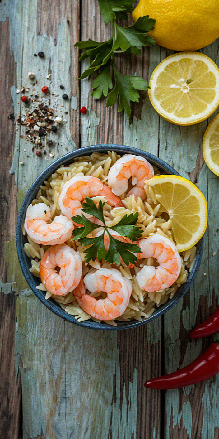 A rustic wooden table with a bowl of shrimp pasta garnished with parsley, surrounded by lemons, chili peppers, and peppercorns.