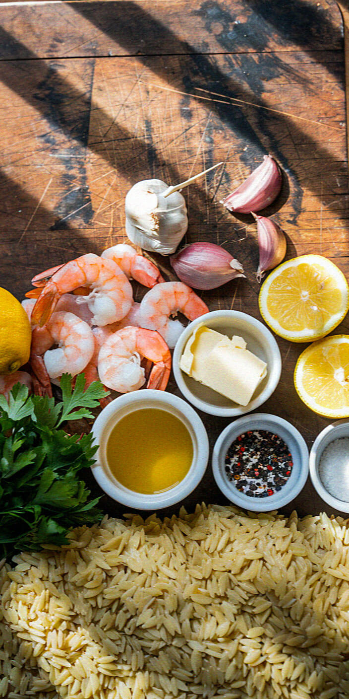 A wooden surface displays shrimp, garlic, and lemon, with bowls of butter, seasoning, and oil next to fresh parsley and uncooked pasta.