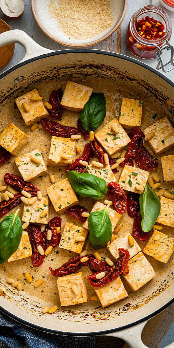 A dish with tofu cubes, sun-dried tomatoes, basil leaves, and pine nuts in a pan on a wooden surface with seasoning jars nearby.
