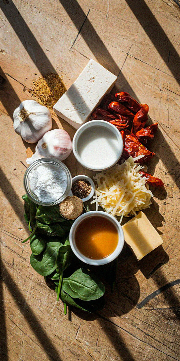 A variety of fresh ingredients including garlic, spinach, and cheese arranged artistically on a wooden surface, illuminated by natural sunlight.