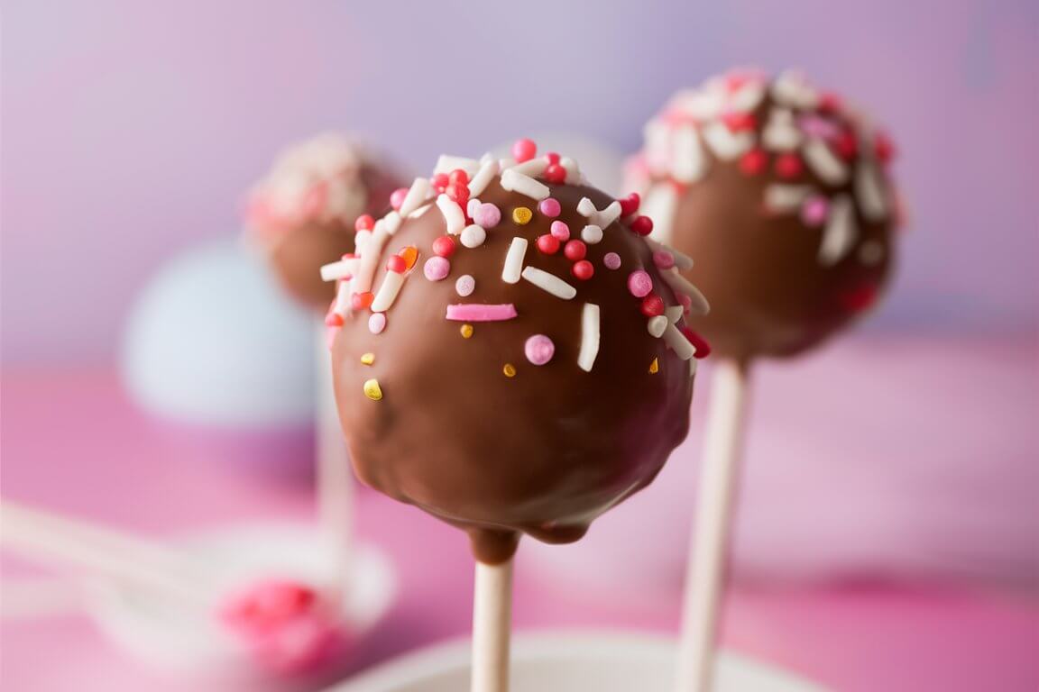 Close-up of chocolate cake pops with colorful sprinkles on sticks, set against a soft pink and purple background.