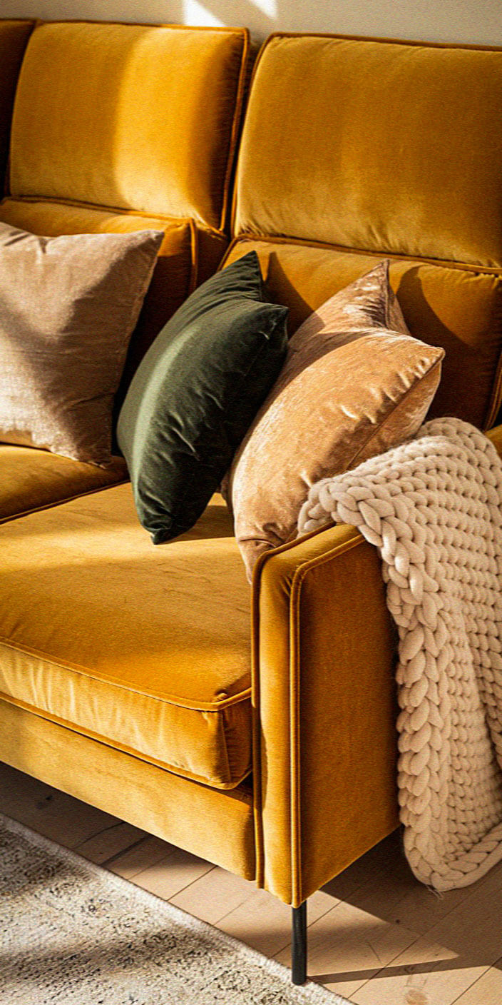 A cozy yellow sofa with green and beige pillows, draped with a chunky knit blanket, illuminated by soft natural light.