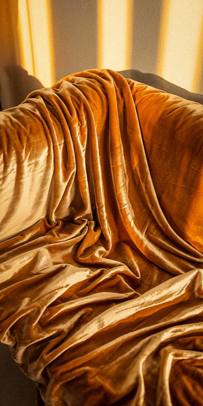 Golden velvet fabric draped over a chair, bathed in warm sunlight, casting soft shadows. The texture appears luxurious and inviting.