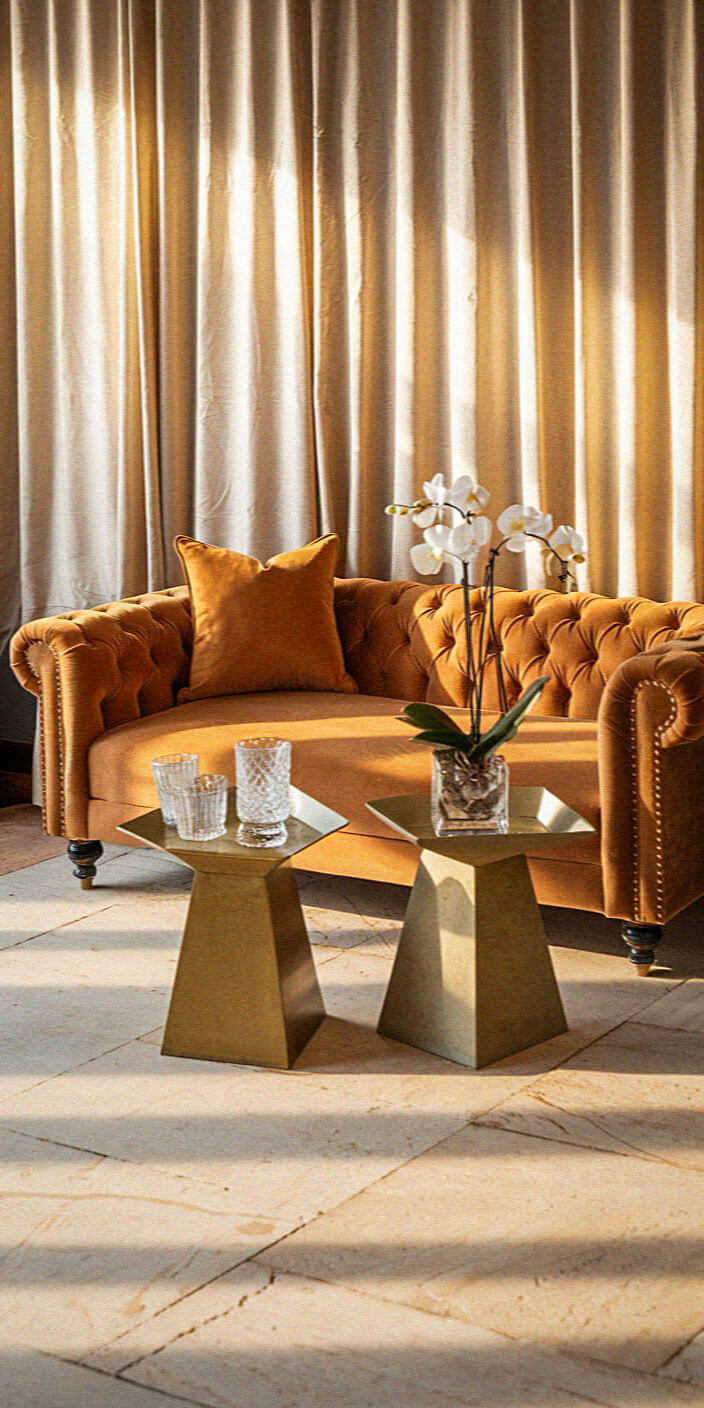A cozy living room with a tufted sofa, velvet pillow, gold tables, glassware, and orchids, framed by elegant beige curtains.