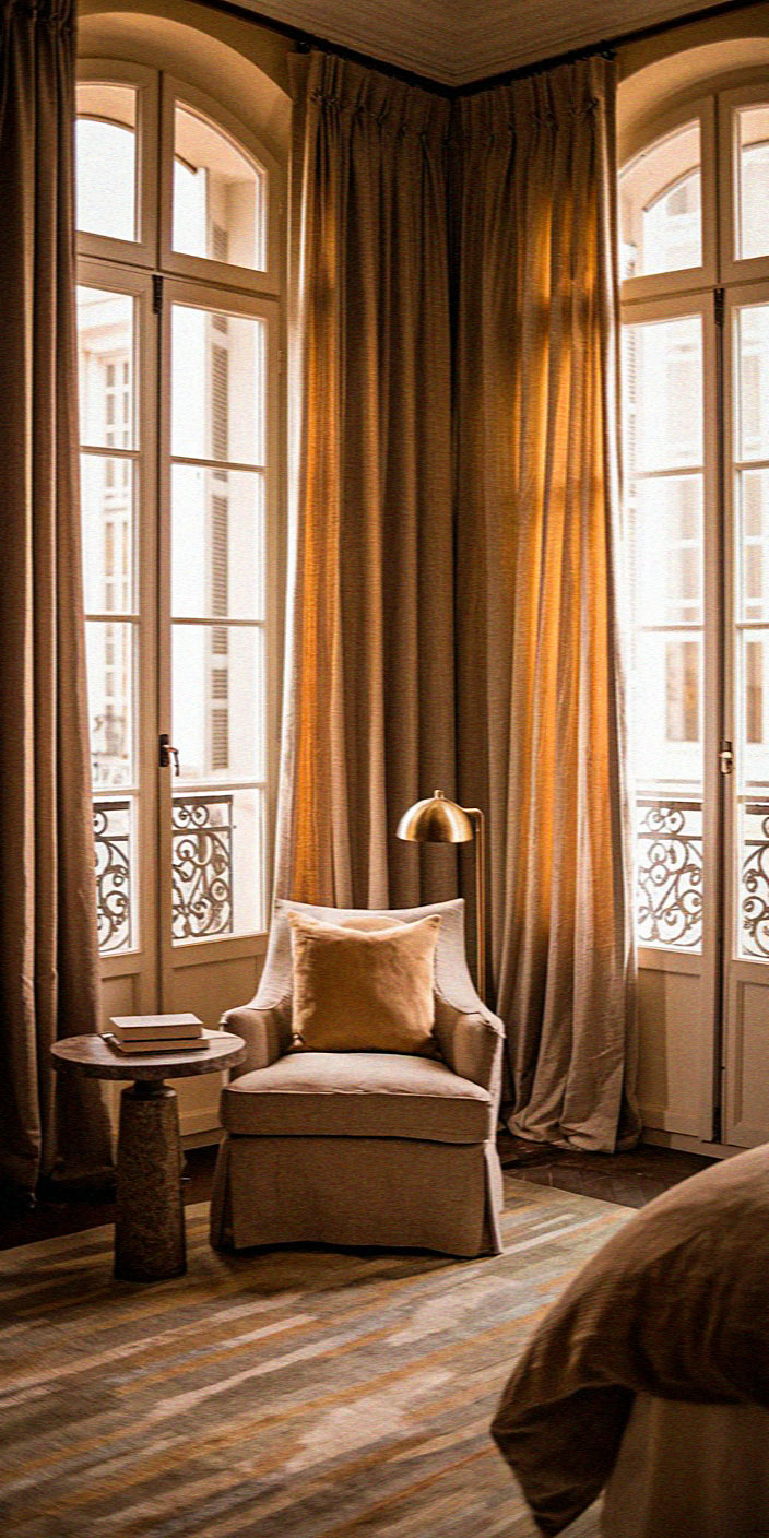 Elegant room with tall arched windows, warm curtains, cozy armchair, small round table, and lamp. Soft natural light creates a serene atmosphere.