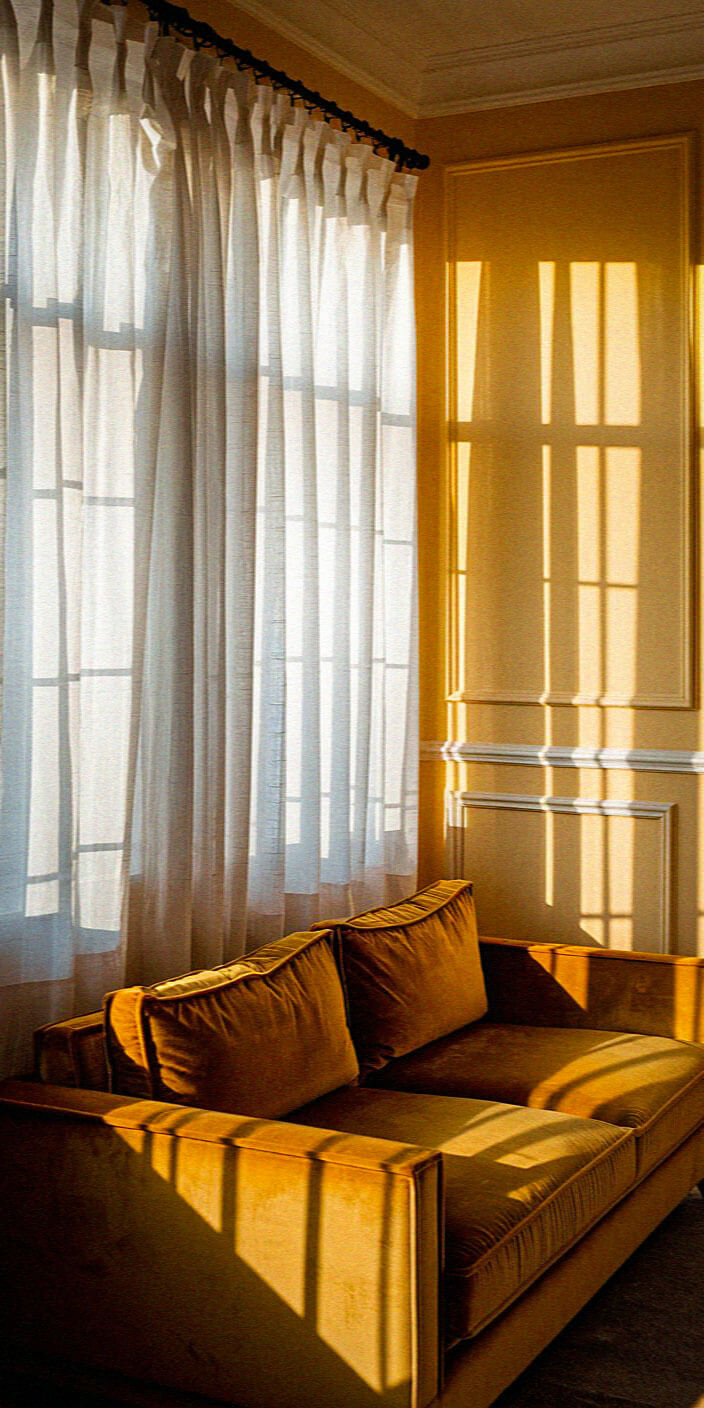 Sunlight streams through sheer curtains, casting shadows on a warm, yellow sofa in a softly lit, elegant room with detailed wall paneling.