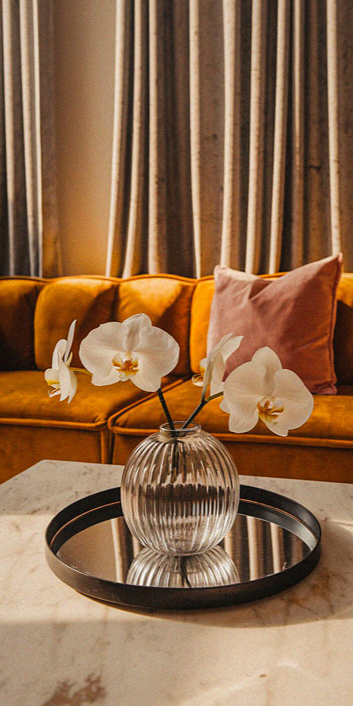 A cozy interior features a marble table, a round glass vase with white orchids, mustard sofa, pink pillow, and elegant curtains.