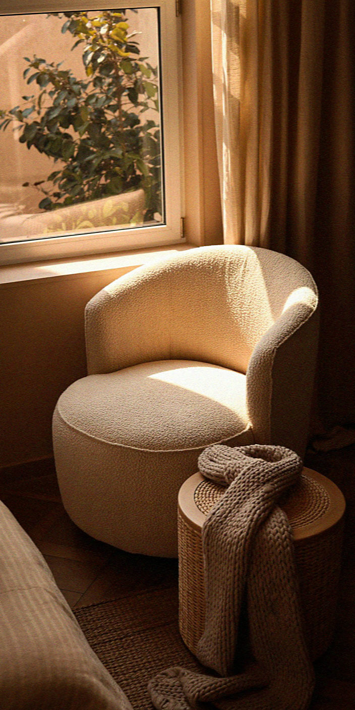 A cozy room features a soft, beige chair beside a window with sunlight streaming in, and a knitted throw on a side table.