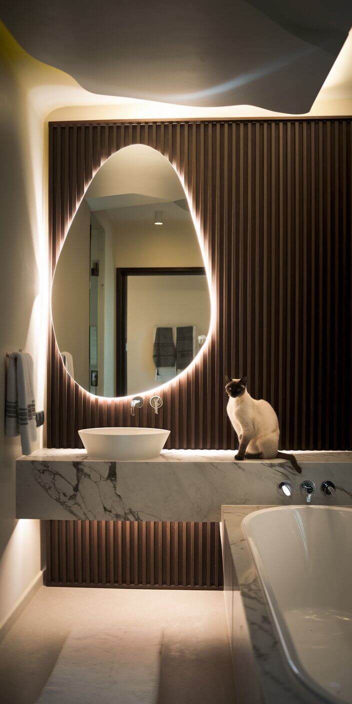 Modern bathroom with marble counter, backlit mirror, and elegant bathtub. A cat sits by the sink, enhancing the serene ambiance.