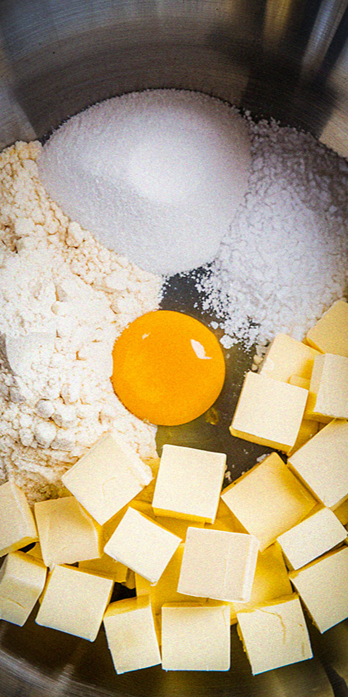 A bowl contains flour, sugar, an egg yolk, and cubed butter, ready for baking preparation in a stainless steel mixing bowl.