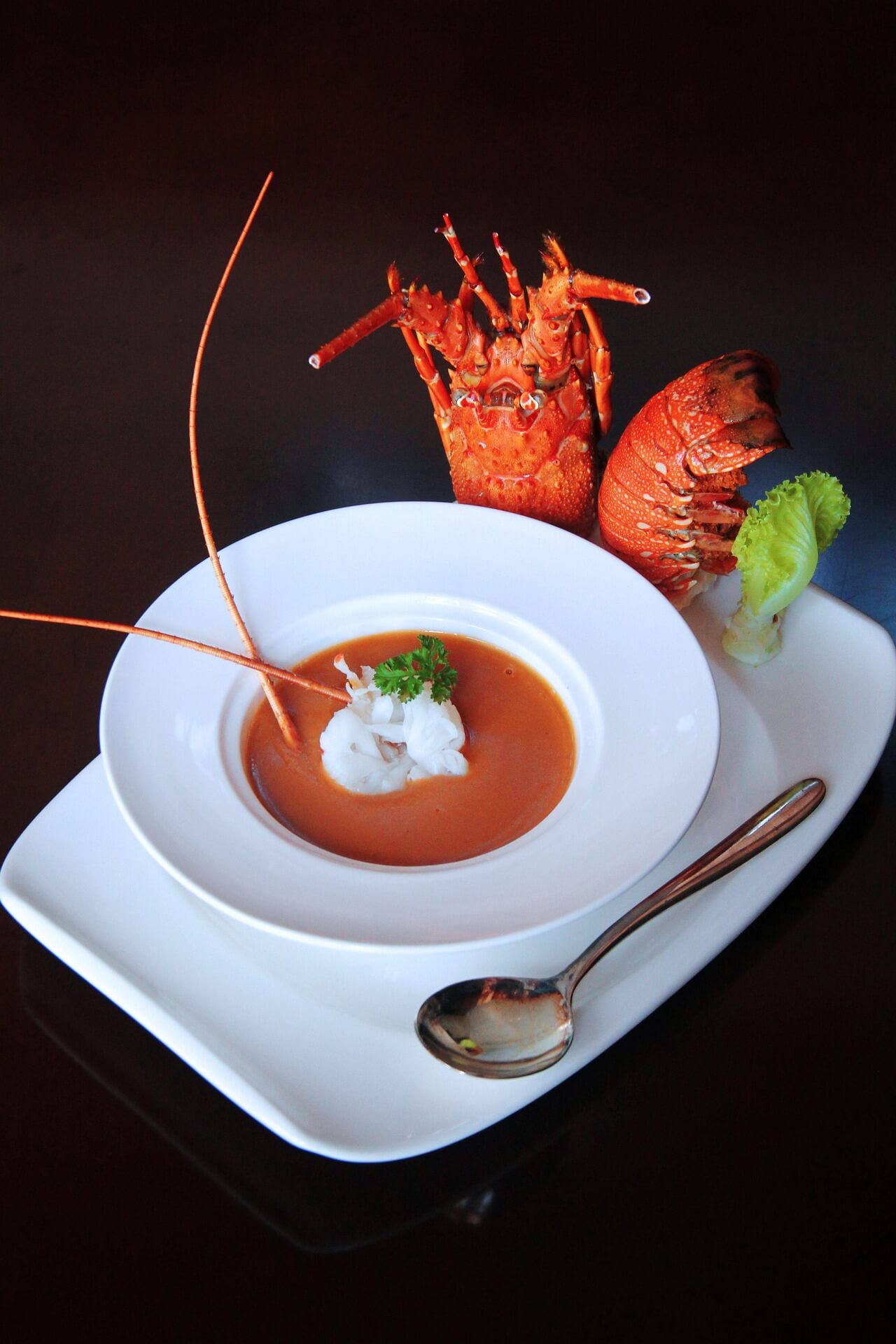 A white bowl of orange lobster bisque topped with a dollop of cream and herbs, set on a white plate with lobster tails visible in the background.