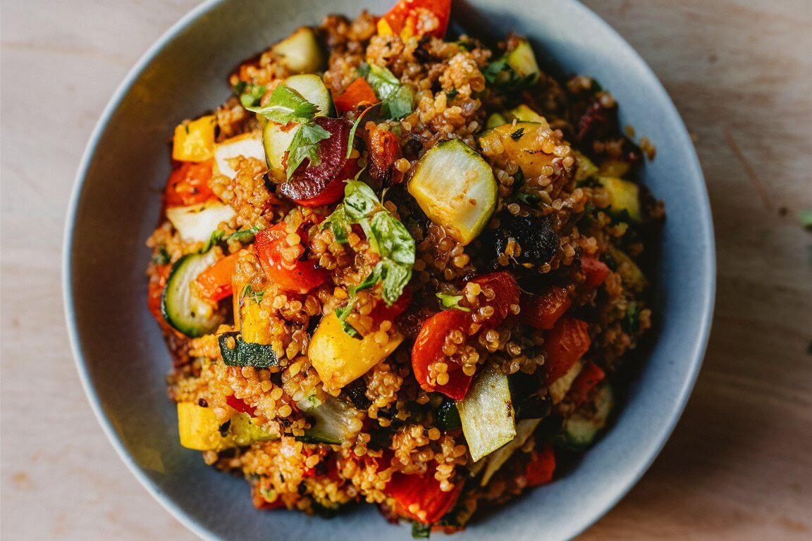 A bowl of colorful roasted vegetable quinoa salad with zucchini, tomatoes, peppers, and fresh herbs sits on a light wooden table.