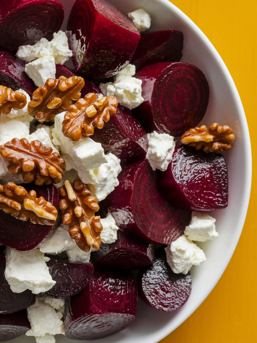 A bowl contains sliced beets, crumbled cheese, and walnuts, set on a vibrant yellow background creating an appealing and colorful dish.