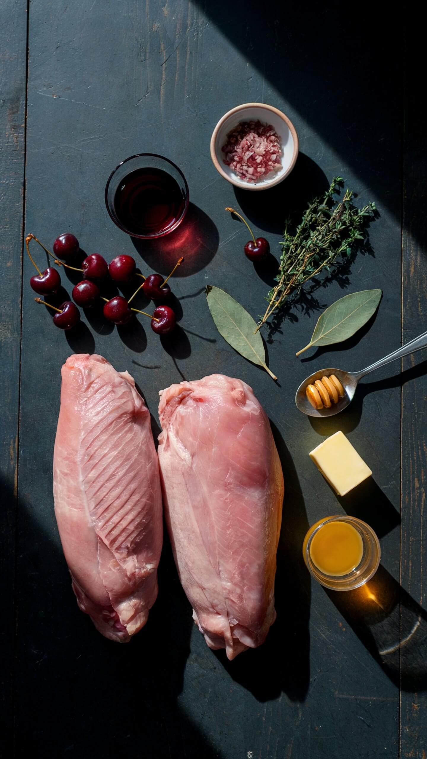 Raw turkey breasts, cherries, bay leaves, thyme, honey, butter, red wine, and chopped onions on a dark surface under partial sunlight.