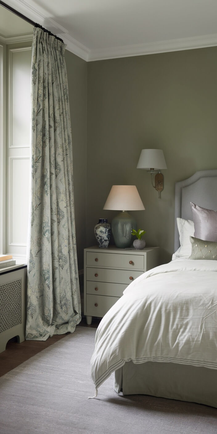 Elegant bedroom with pale green walls, floral curtains, and a cushioned bed. Bedside table holds two lamps and decorative items.