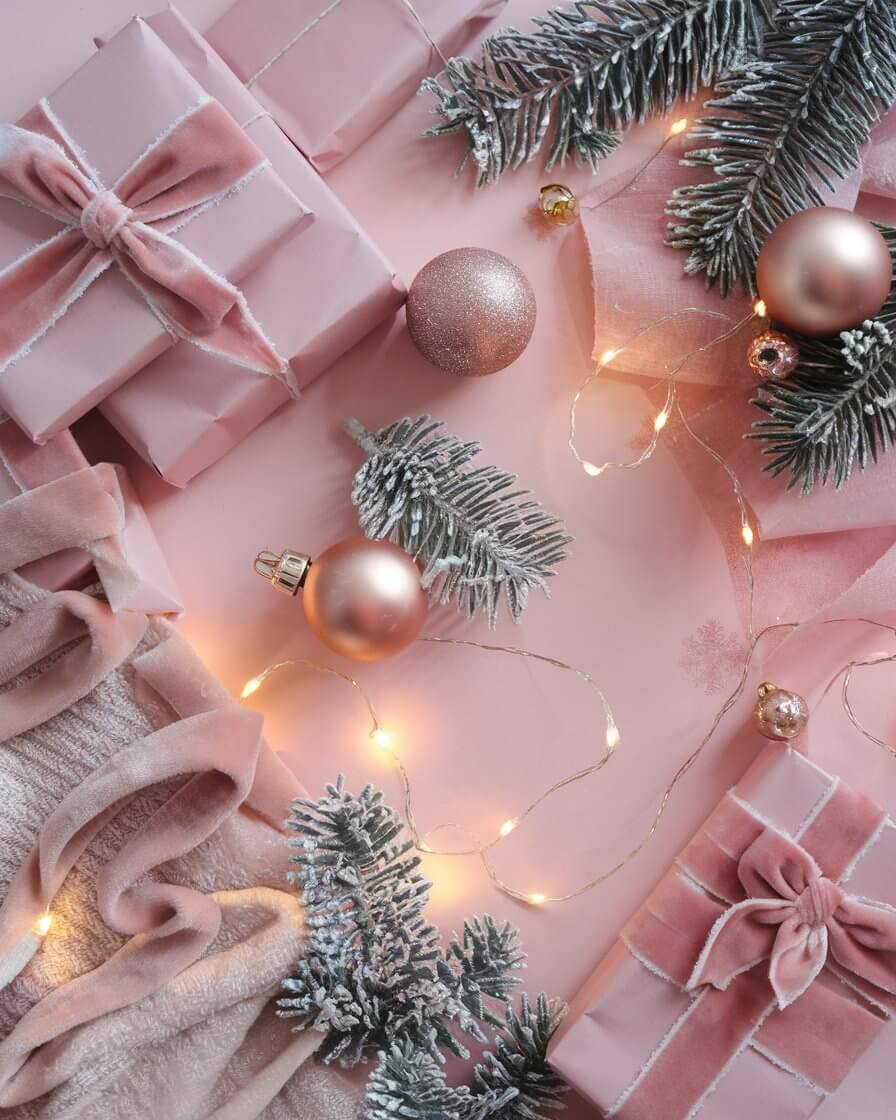 Pink gift boxes with velvet ribbons, surrounded by frosted pine branches, ornaments, and glowing fairy lights on a soft pink background.