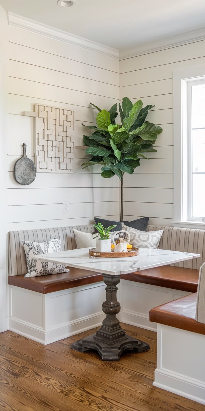 A cozy breakfast nook features white shiplap walls, a potted plant, patterned pillows, and a decorative table with a tray holding small items.