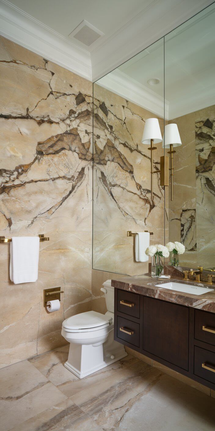 Elegant bathroom with marble walls, large mirror, modern fixtures. Features a vase of white flowers, towels, and dark wood vanity with gold accents.