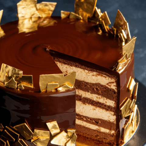 Elegant chocolate cake with glossy glaze and gold leaf decorations. A slice is cut, revealing layered interior on a dark backdrop.