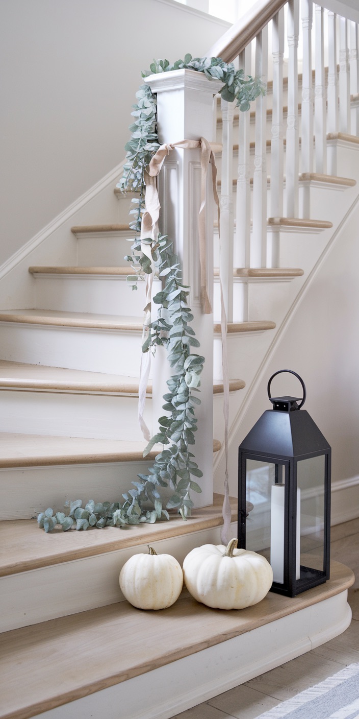 White pumpkins and a black lantern decorate a staircase with eucalyptus garland, creating a simple and elegant seasonal arrangement.