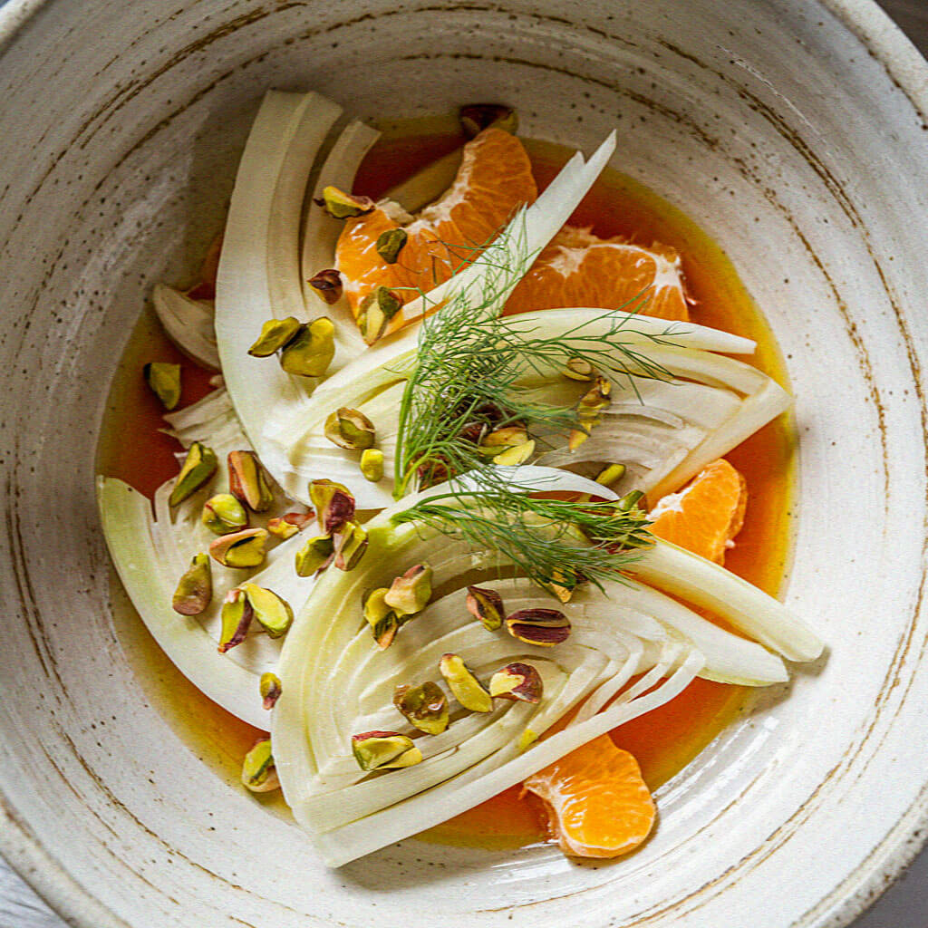 A ceramic bowl holds fennel slices, orange segments, pistachios, and a sprig of fennel, arranged in vibrant citrus dressing.