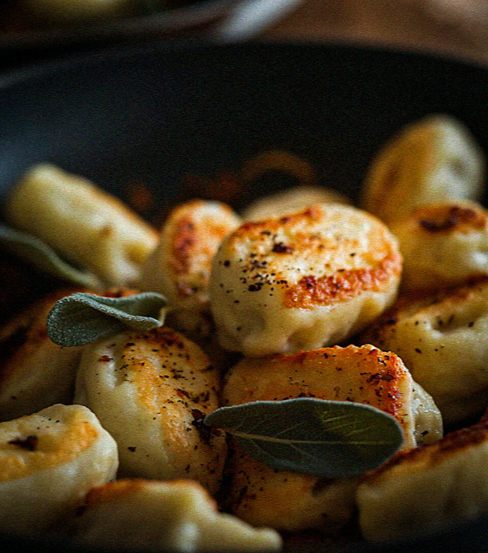 Golden-brown dumplings garnished with fresh sage leaves in a dark, shallow pan, creating a warm, appetizing visual focused on rustic culinary presentation.