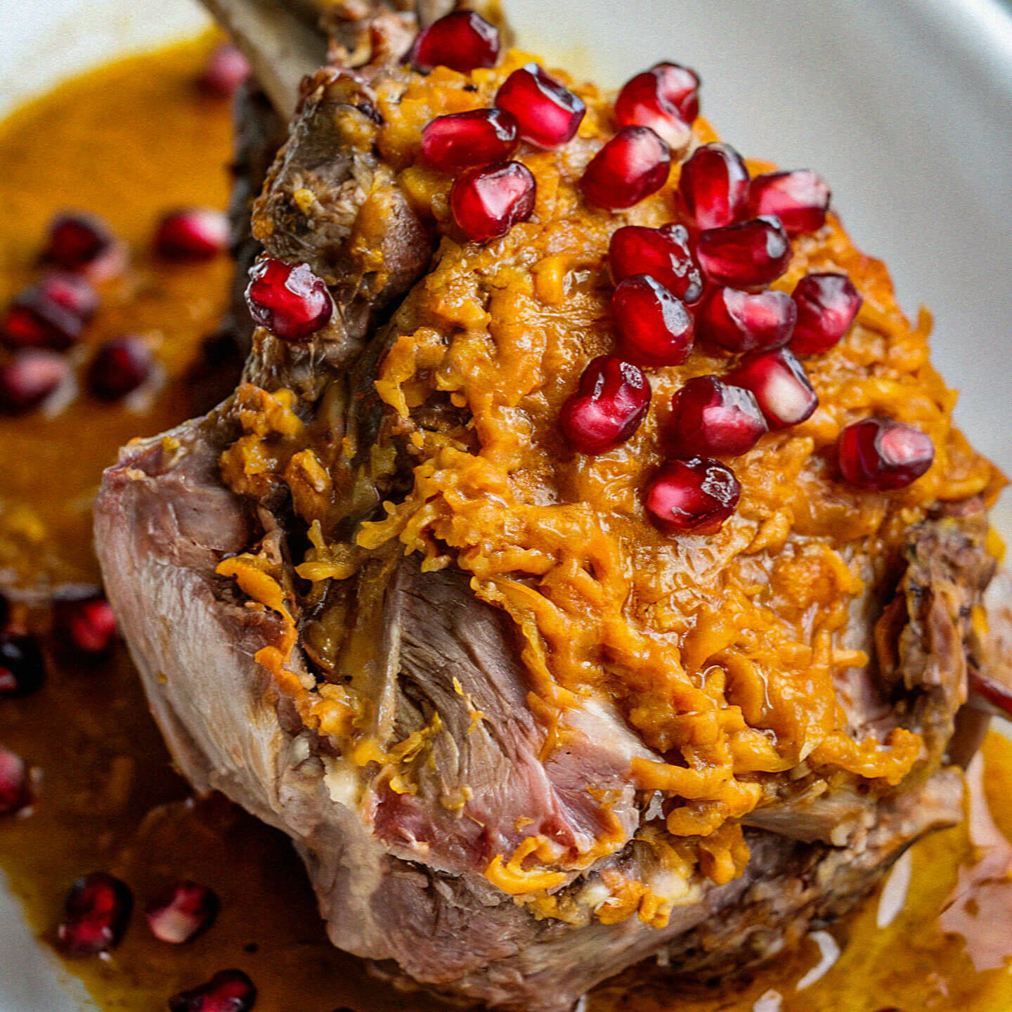 A deliciously roasted lamb shank topped with pomegranate seeds and orange zest, served on a white plate with savory sauce.