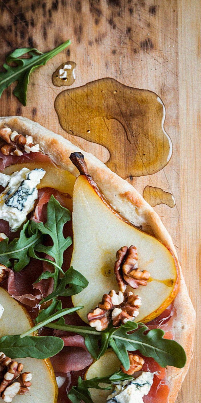 Gourmet pizza on wooden board topped with pear slices, walnuts, arugula, blue cheese, and prosciutto. Honey drizzle adds sweetness beside it.