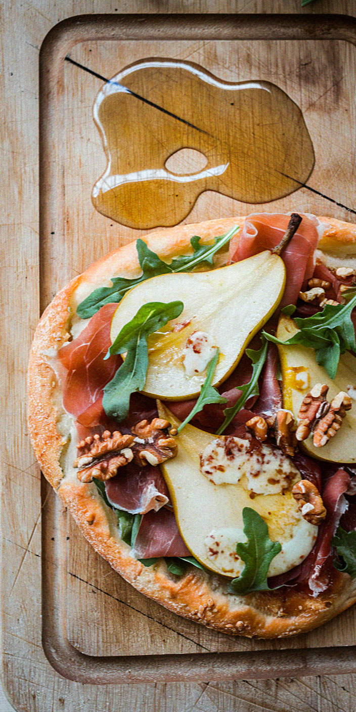 A rustic pizza topped with pear slices, arugula, prosciutto, walnuts, and cheese sits on a wooden board with a splash of honey.
