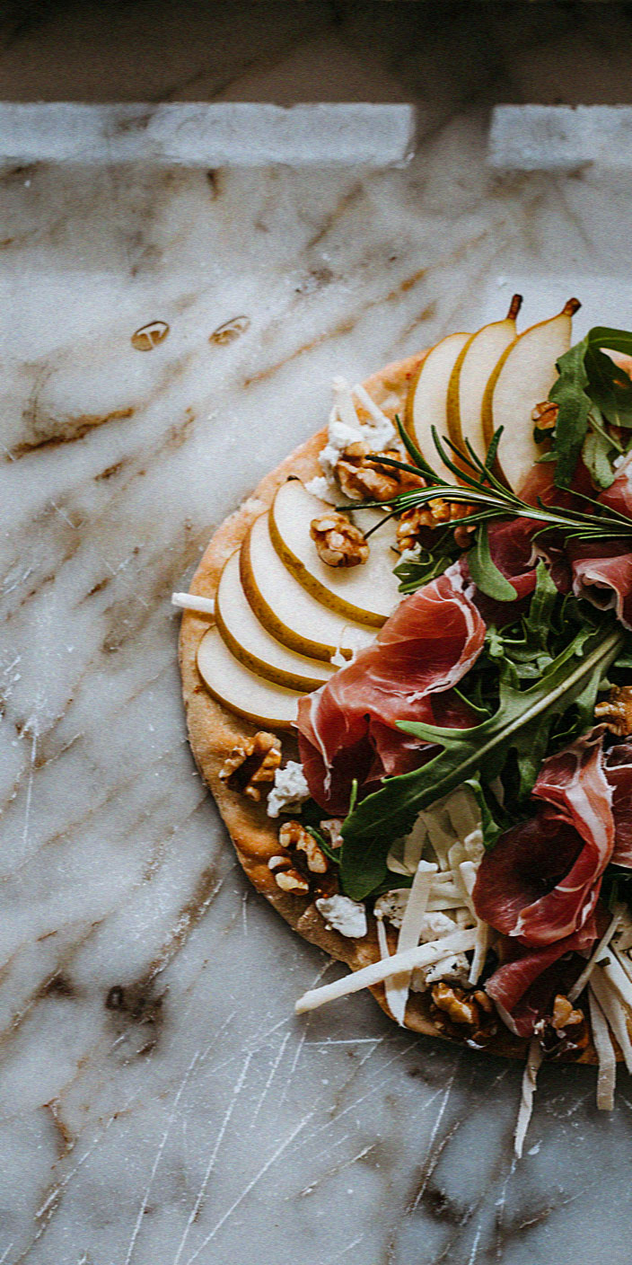 A rustic pizza topped with pear slices, prosciutto, arugula, walnuts, and cheese on a marble surface, creating an artisanal culinary presentation.